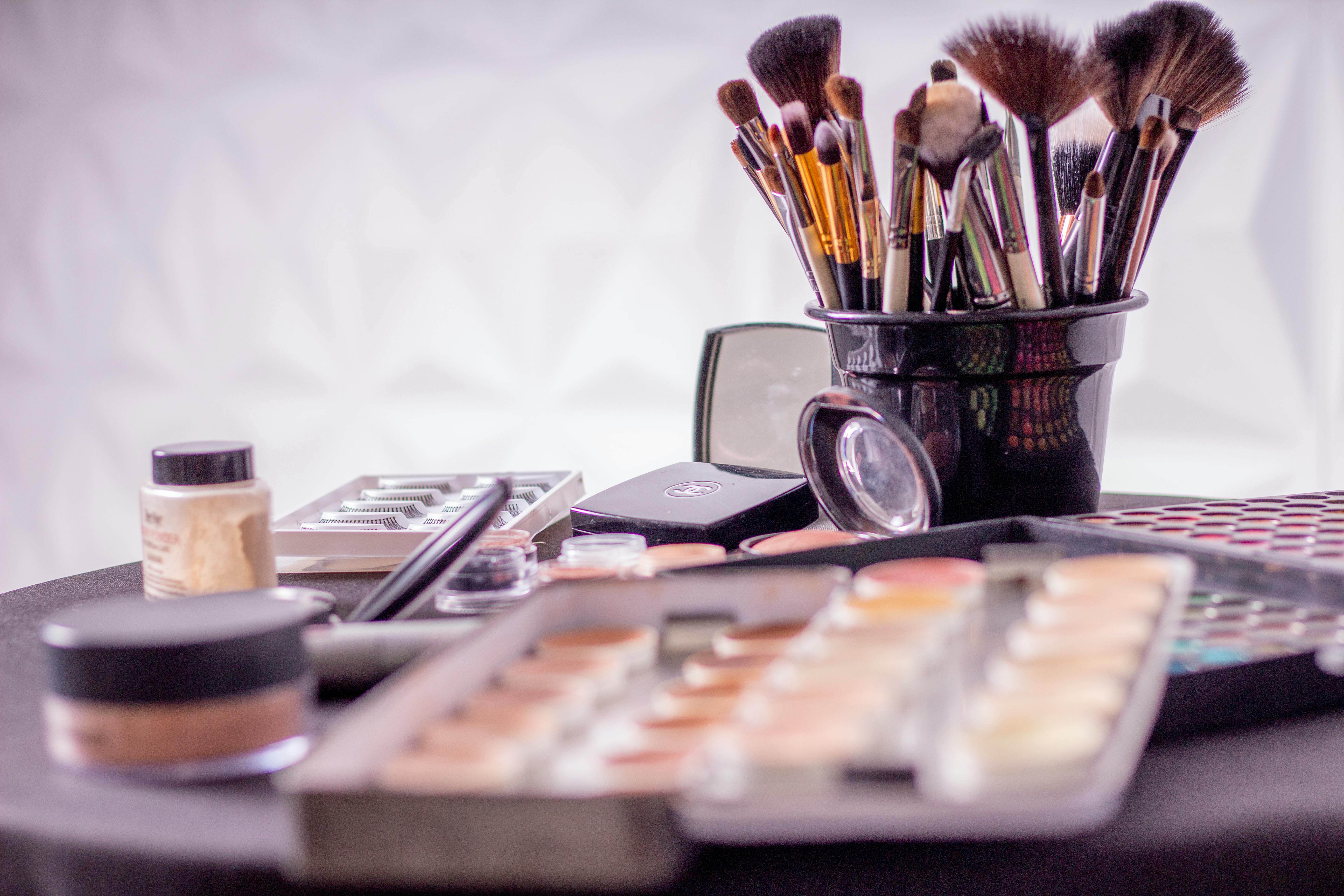 A cup of makeup brushes with eyeshadow and various cosmetics on a table.